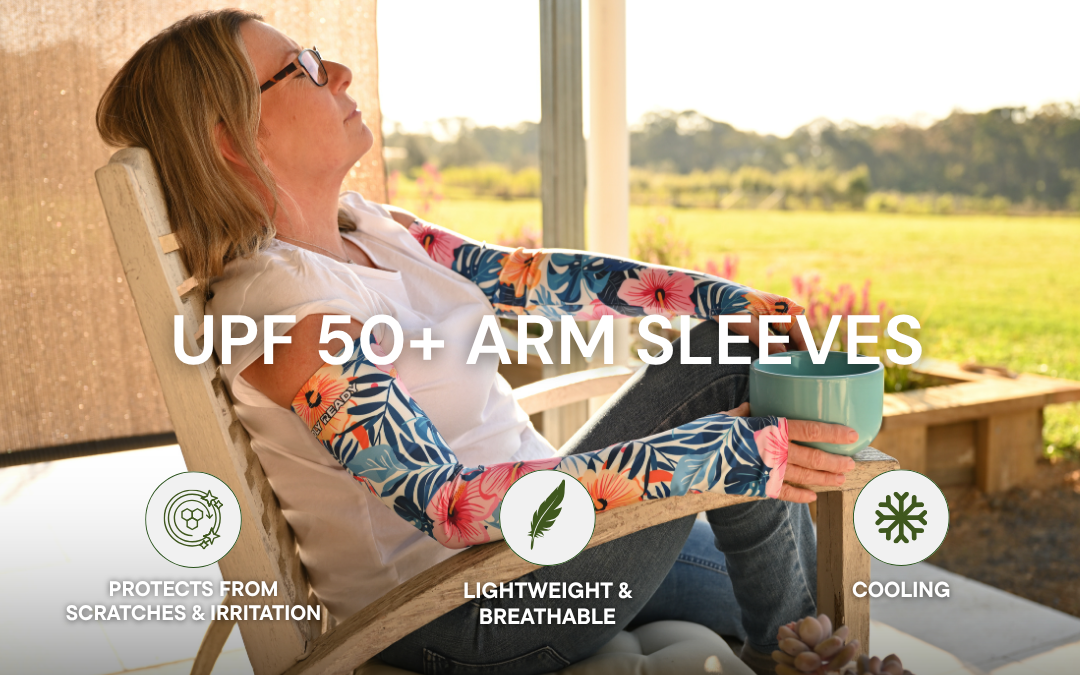 A woman reclining in a wooden chair on a porch wearing colorful floral arm sleeves with a tropical leaf pattern. Large text reads 'UPF 50+ ARM SLEEVES' with feature icons below for scratch protection, lightweight breathability, and cooling.